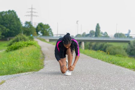 African woman pausing to tie her shoe laces while out jogging along a rural road alongside a river in a health and fitness conceptの写真素材