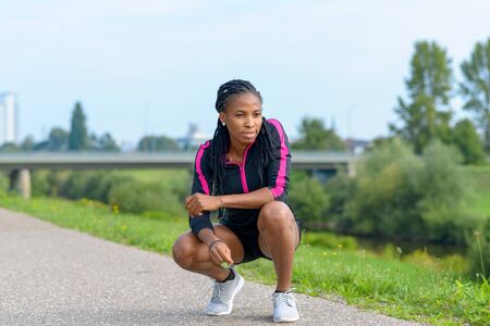 Fit sporty young African woman crouching on a rural road looking ahead with a thoughtful expression as she takes a rest during her daily jogging workout in a health and fitness conceptの写真素材