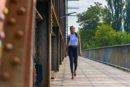 Trendy athletic young African woman laughing as she approaches the camera along an old wooden pedestrian bridge in a low angle viewの写真素材
