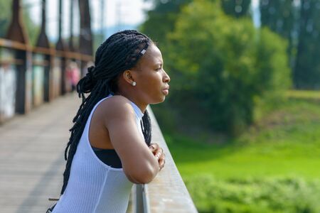 Young African woman standing leaning on the wooden railing staring off a bridge deep in thought in a profile viewの写真素材