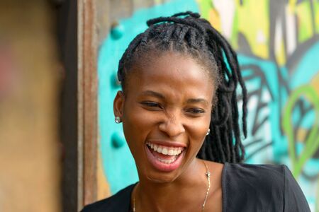 Attractive young African woman with a vivacious smile laughing at the camera as she poses in front of colorful graffitiの写真素材