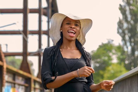 Laughing vivacious young African woman wearing braided hair extensions and a stylish wide brimmed straw hat in a close up outdoor portraitの写真素材