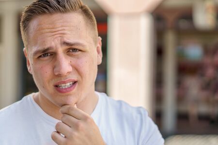 Thoughtful troubled young man with his hand to his chin frowning at camera outdoors in an urban streetの写真素材