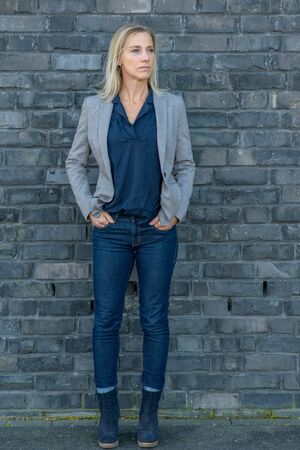 Full length shot of an attractive middle-aged fit woman with long blond hair looking away while wearing cool slim blue jeans, T-shirt and boots with a gray jacket outdoorsの写真素材