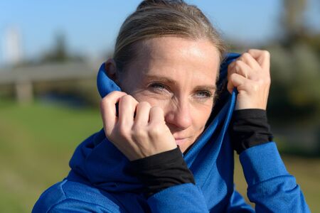 Attractive stylish blond woman in blue outfit pulling the hood of her top over her head with a...