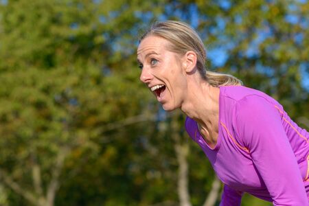 Playful vivacious blond woman laughing as she leans forwards striking a pose outdoors against a...