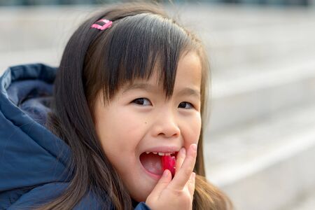 Pretty little Asian girl popping a colorful red sweet into her mouth while glancing sideways at the...