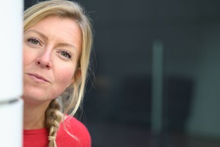 Pretty middle-aged blond woman with pensive look peering at the camera around a white pillar in a close up portraitの写真素材