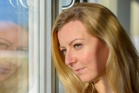 Attractive blond woman standing daydreaming looking longingly out through a window reflected in the...