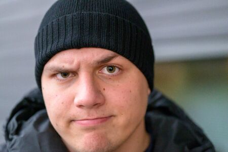 Determined sceptical young man wearing a beanie and warm jacket standing outdoors in winter frowning...