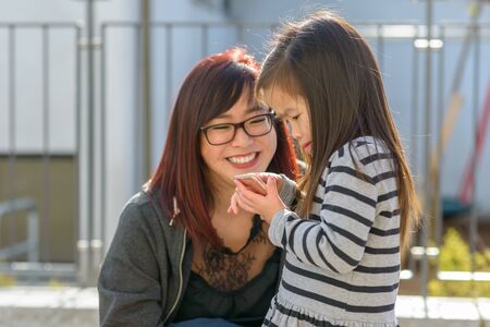 Small little girl playing with a mobile phone held in a loving embrace by her smiling happy young...