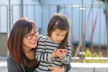 Small little girl playing with a mobile phone held in a loving embrace by her smiling happy young...