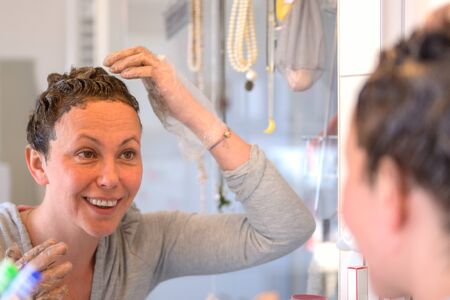 Smiling Hispanic woman applying hair dye to her curly hair at home with an over the shoulder view to her reflection in the mirrorの写真素材