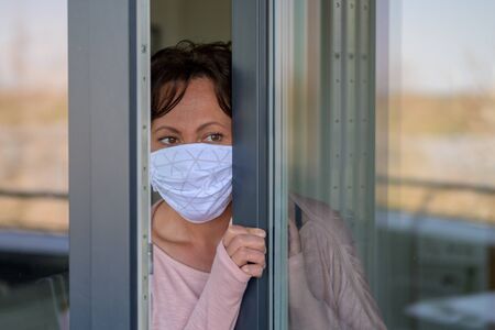 Woman wearing a face mask peering around anxiously as she looks through a partly opened glass window...