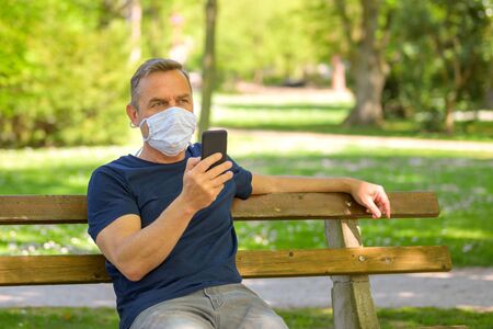 Man wearing a face mask sitting using a mobile phone outdoors on a bench in a lush green park in spring in a concept of the coronavirus or Covid-19 pandemicの写真素材