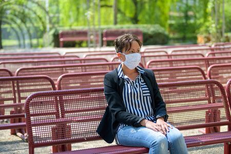 Woman seated on empty benches outdoors in a park wearing a surgical mask as infection control during the coronavirus or Covid-19 pandemic, with lateral copy spaceの写真素材