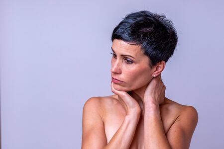 Thoughtful attractive woman with pensive expression and bare shoulders holding her hand to her throat as she stares upwards over grey with copyspaceの写真素材