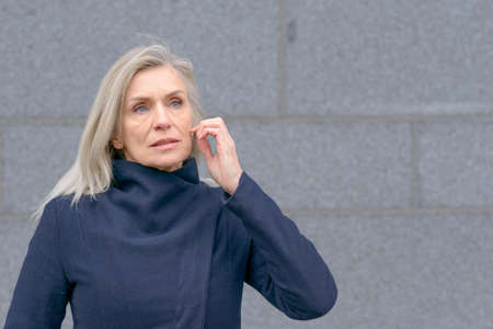 Attractive middle-aged blond woman with a serious worried expression looking up listening against a grey exterior wall with copyspaceの写真素材