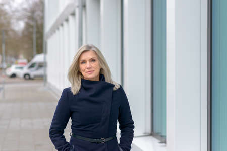 Attractive blond woman in a stylish blue coat standing in a city street in winter looking thoughtfully at the camera with copyspaceの写真素材