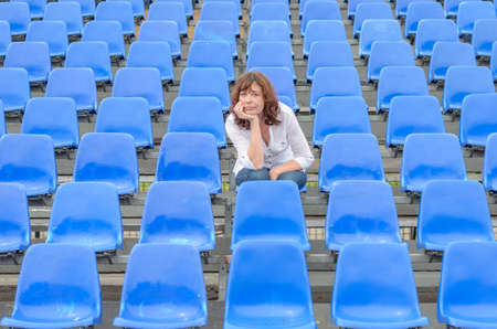 Glum woman sitting in empty rows of blue spectator seating in an auditorium or stadium with a bored expression and her chin on her handsの写真素材