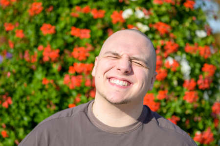 Candid portrait of a young man screwing his eyes shut while smiling outdoors in the sunshine against a background of colorful red flowers in the gardenの写真素材