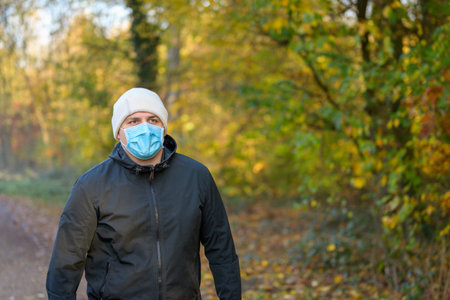 Young man hiking in woodland in autumn a protective face mask during the coronavirus or Covid-19 pandemic approaching the camera with copyspaceの写真素材