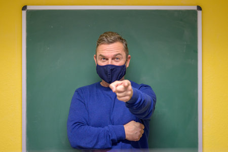 Male teacher wearing a cloth face mask during   pandemic pointing at the camera with raised eyebrows...