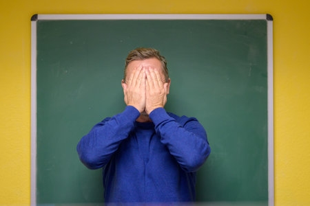 Male teacher covering his eyes with his hands as he stands in front of a wall mounted chalkboard in a classroomの写真素材