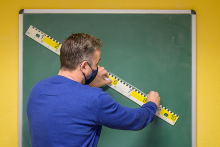 Back view of a teacher using a ruler to draw a straight diagonal line on a chalkboard while wearing...