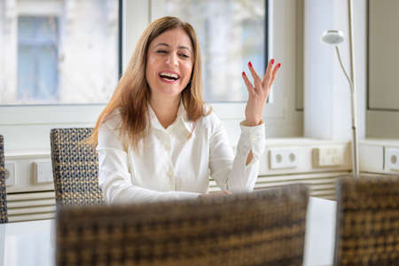Vivacious animated woman gesturing with her hand and laughing as she sits at a conference table in a high key office or apartment with window backdropの写真素材