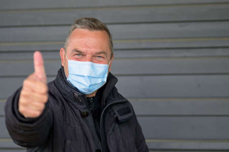 Smiling middle-aged man with laughing eyes wearing a face mask as he poses outdoors against a grey wall during the  pandemic giving a thumbs up sign of approval or to show all is goodの写真素材