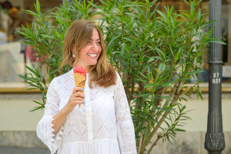 Stylish woman holding a berry ice cream cone in her hand as she stands in front og a green bush...