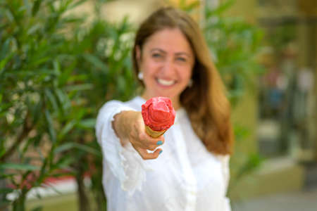 Happy smiling woman holding a cone of berry ice cream extended to the camera with focus to the...