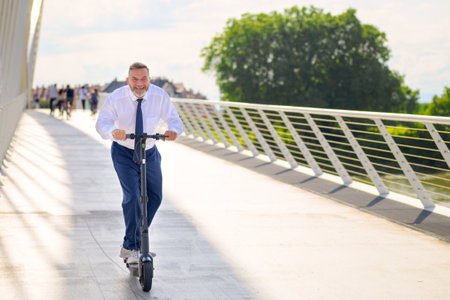 Gleeful businessman enjoying his ride on an electric scooter as he crosses a long bridge at sunset using the pedestrian walkway in an urban transport conceptの写真素材