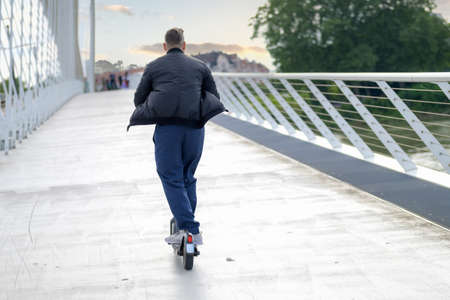 Rear view of a man riding an electric scooter on a bridge at sunset towards distant people crossing the walkway in an urban transport conceptの写真素材