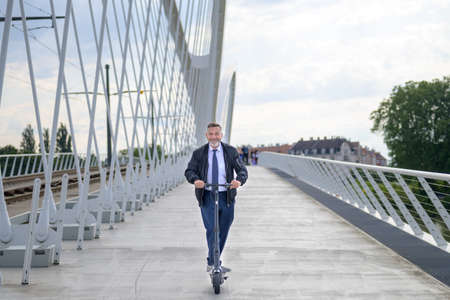 Businessman commuting in the city using an electric scooter smiling as he approaches the camera while crossing a bridge over a river at sunsetの写真素材