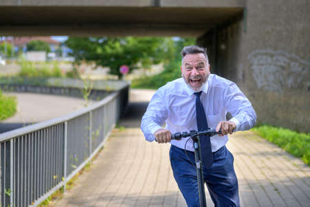 Jovial businessman yelling in pure enjoyment as he speeds along on an electric scooter approaching...