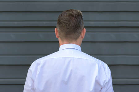 Rear head and shoulders view of a man in white long sleeved shirt standing facing a grey exterior...