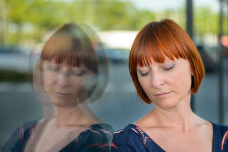 Dejected woman with a sad expression and downcast eyes standing leaning against a store window in town reflected in the glass in a close up headshotの写真素材