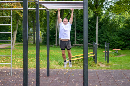 Middle-aged man having fun working out in a park hanging from a set of bars by his arms at an...