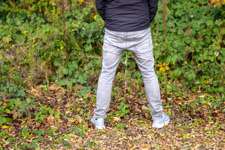 Rear view of the lower body of a man in jeans and sneakers urinating outdoors in a park or garden