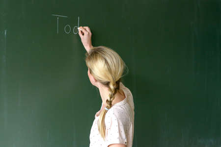 Female teacher with blond hair ponytail writing with chalk on top of a clean green chalkboard