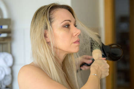 Woman drying and styling her long blond hair after washing it with an electric hairdryer and circular brushの写真素材