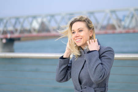 Attractive woman standing on a waterfront promenade on a windy day clutching at her long blond hair with a smileの写真素材