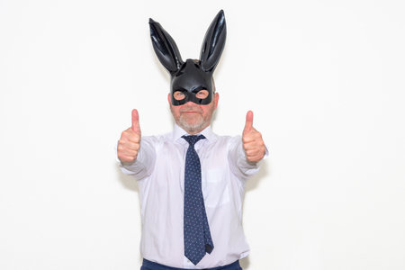 Businessman wearing a bunny mask giving a thumbs up with both hands as he stands smiling at the camera over whiteの写真素材