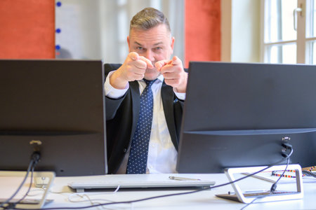 Businessman pointing with both hands at the camera viewed between computer monitorsの写真素材