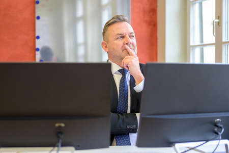 Businessman sitting pondering a problem in the office with hand to chin as he stares asideの写真素材