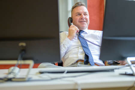 Confident businessman talking on a telephone in an office as he relaxes sitting slumped back in his chairの写真素材
