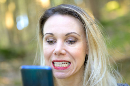 Attractive blond woman examining her teeth in a hand mirror outdoors in a close up head shot