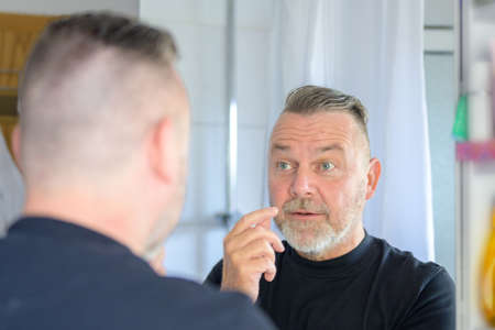 Dismayed middle-aged man looking at the fleshy bags under his eyes with a view to his reflection in...